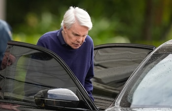Un hombre de cabello blanco y suéter azul oscuro se inclina para ingresar a un automóvil negro con la puerta abierta