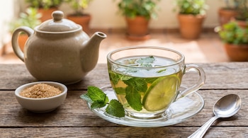 Una taza de té de menta con lima y hojas, un platillo, una tetera, un cuenco con azúcar moreno y una cuchara sobre una mesa de madera.