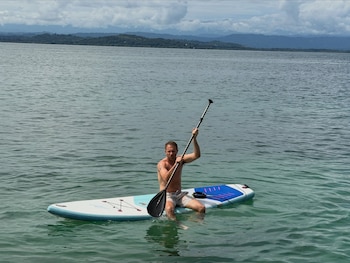 Un hombre sentado en una tabla de paddle surf sostiene un remo en el agua, con una costa montañosa y nubes de fondo