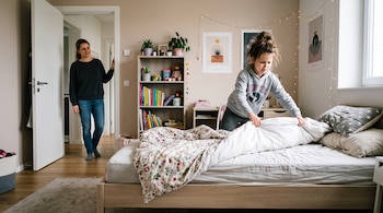 Una niña acomoda las sábanas de una cama de madera mientras una mujer la observa desde el marco de la puerta.