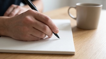 Primer plano de una mano izquierda que escribe con un bolígrafo negro sobre un cuaderno blanco. Hay una taza beige desenfocada en una mesa de madera.