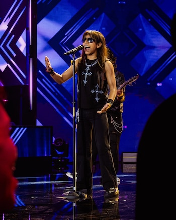 Una persona joven canta frente a un micrófono sobre un escenario con luces azules, vestida de negro y con gafas oscuras