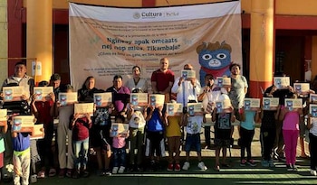 Grupo de adultos y niños sonrientes sosteniendo certificados en un evento al aire libre; detrás, un gran cartel con el logo de Cultura e INALI y un dibujo de un gato azul