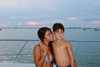 Mujer de cabello oscuro con bikini azul y niño sin camiseta posan en un barco. En el fondo se ven el mar, un cielo nublado, edificios y veleros