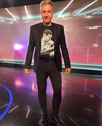 Un hombre de mediana edad, con cabello gris, viste un blazer negro sobre una camiseta negra con una imagen de Lionel Messi, de pie en un estudio de televisión con luces