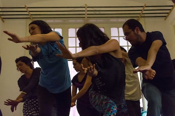 Cinco personas realizan un movimiento de danza con los brazos extendidos y las rodillas flexionadas en una sala con paredes claras, ventanas y vigas