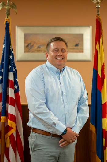 Un hombre con camisa clara posa sonriendo entre la bandera de Estados Unidos y la de Arizona, con un cuadro en la pared