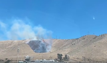 Vista panorámica de un incendio forestal activo en una colina árida. Grandes llamas y humo blanco-grisáceo cubren la ladera, con torres eléctricas en la cima y un cielo azul claro