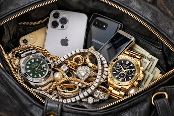 Primer plano de un bolso de cuero negro abierto mostrando celulares, relojes de lujo, joyas de oro con diamantes y perlas, y billetes de dólar.