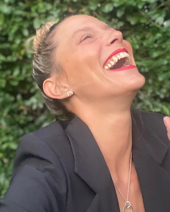 Primer plano de Manuela Pal riendo a carcajadas con la cabeza hacia atrás, labios rojos, blazer oscuro y collar plateado, sobre fondo verde de hojas