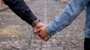 Un hombre y una mujer tomados de la mano mientras caminan por una calle empedrada; el hombre lleva una camisa oscura y reloj, la mujer una chaqueta vaquera.