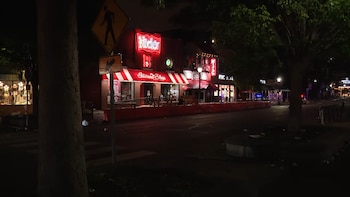 Vista nocturna de una calle con tiendas iluminadas por letreros de neón, incluyendo 'Nick's' y 'The Indiana Shop', con barreras rojas y algunos desechos