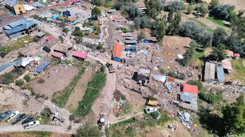 Vista aérea de un pueblo con numerosas casas destruidas y escombros. Se observan personas, vehículos, campos de cultivo y árboles