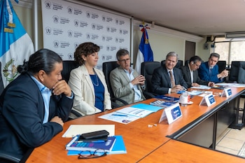 Vista de un grupo de seis personas sentadas alrededor de una mesa de conferencias de madera en una sala. Hay banderas y un logo institucional en el fondo
