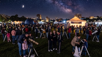 Personas agrupadas sobre el césped observan el cielo con telescopios junto a carpas iluminadas durante la noche.