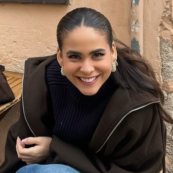 Una mujer sonriente con cabello recogido, chaqueta marrón, suéter oscuro y pendientes blancos, sobre un fondo de pared beige