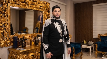 Hombre con chaqueta negra bordada y capa junto a una consola dorada con botellas de licor y un espejo ornamentado.