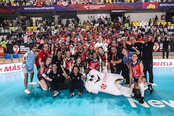 Un equipo de voleibol femenino y su personal celebran en la cancha de un estadio lleno, posando junto a una mascota de diente gigante. Al fondo, la grada con aficionados