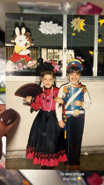 Fotografía retro de una niña vestida con traje de época rojo y negro con abanico, junto a un niño con uniforme de soldado y bigote pintado. Ambos sonríen