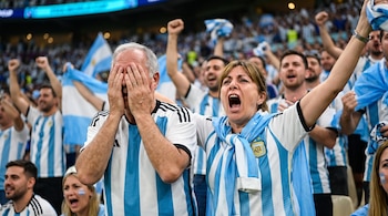 Hombre con camiseta de Argentina cubriendo su rostro. Mujer con camiseta de Argentina gritando. Ambos en primer plano, rodeados de hinchas borrosos en el estadio.