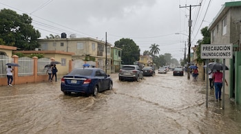 Calle inundada por lluvia en República Dominicana. Coches avanzan y personas caminan con paraguas. Un cartel indica "PRECAUCIÓN INUNDACIONES".