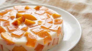 Gelatina de mosaico con cubos anaranjados y trozos de durazno en base blanca. Se presenta en un plato blanco con un fondo claro y desenfocado.