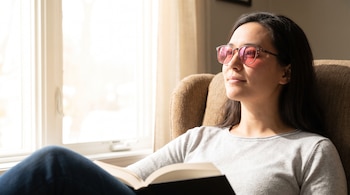 Una mujer sentada en un sillón junto a una ventana lleva anteojos de marco oscuro con cristales de tinte rosado y sostiene un libro abierto.