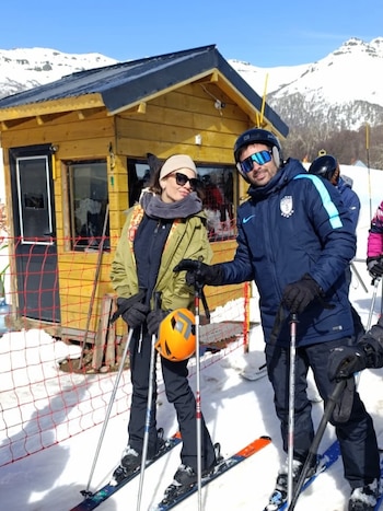 Un hombre y una mujer con ropa de esquí y bastones posan sobre la nieve frente a una casilla de madera en un paisaje montañoso