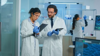 Dos científicos con batas blancas, gafas y guantes azules, uno con tableta y el otro escribiendo, en un laboratorio moderno. Una colega trabaja al fondo.