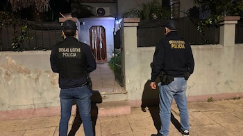 Dos agentes de policía de espaldas con la inscripción Drogas Ilícitas Policía en sus camperas observan la entrada abierta de una casa de noche