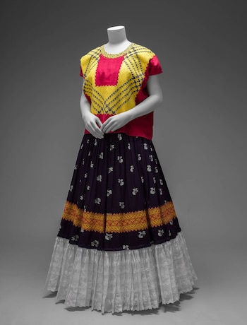 Maniquí blanco de cuerpo completo con un atuendo mexicano: blusa blanca bordada, falda larga negra, y un rebozo rojo con patrones geométricos y franjas púrpuras y doradas
