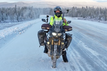 Primer plano de un motociclista en casco y chaqueta verde brillante sobre una motocicleta negra, parado en una carretera nevada con un bosque de pinos y montañas al fondo