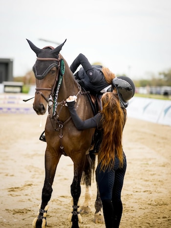 Una mujer con casco se inclina sobre un caballo marrón, mientras otra mujer de pelo largo la abraza por la cintura, en una pista de arena de equitación