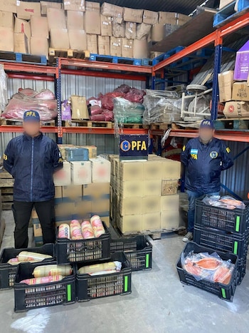 Dos agentes de la PFA con uniforme azul de pie en un depósito. Detrás y en el suelo, cajas apiladas y cajones negros llenos de embutidos y pescado empaquetado