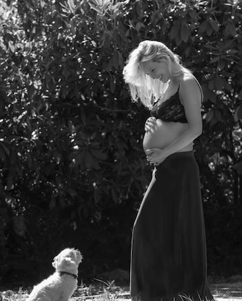 Mujer embarazada rubia sonríe con las manos sobre su vientre. Viste sostén y falda larga. Un perro blanco pequeño mira hacia arriba en un jardín con arbustos frondosos