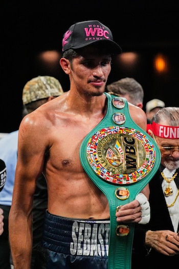 Un boxeador con gorra negra y el torso descubierto sostiene un cinturón deportivo verde y dorado con banderas frente a otras personas