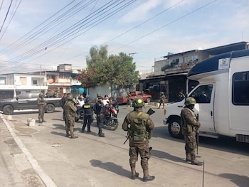 Soldados uniformados con fusiles y agentes de policía inspeccionan a personas en una calle con vehículos oficiales estacionados