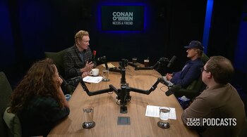 Cuatro personas sentadas alrededor de una mesa de madera con micrófonos en un estudio de pódcast con una pantalla al fondo