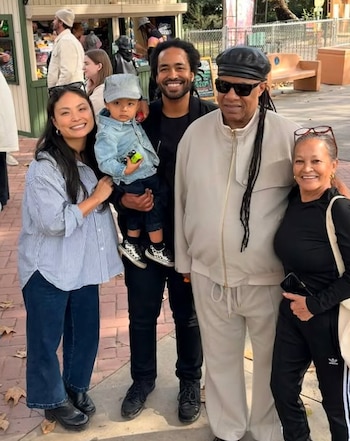Stevie Wonder con gafas de sol, un hombre sosteniendo a un bebé, y dos mujeres sonríen a la cámara en un entorno exterior con edificios y vegetación