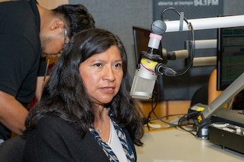 Mujer con cabello oscuro y blusa estampada habla frente a un micrófono en un estudio de radio con equipo técnico y un hombre en segundo plano