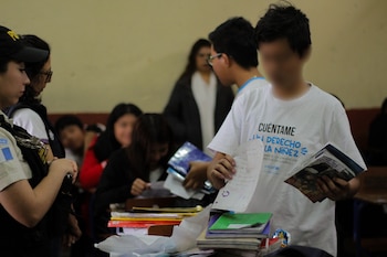 Un joven estudiante con camiseta de UNICEF sostiene papeles y libros. Hay una mesa con cuadernos y otros estudiantes en el fondo, incluyendo una persona con uniforme y gorra