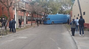 Un bus azul grande bloquea parcialmente una calle urbana. Varias personas, incluyendo personal de seguridad y periodistas, se ven en las aceras adyacentes a edificios.