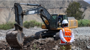 Hombre de espaldas con casco blanco y chaleco naranja de 'Vivienda Equipo de Emergencias' observa una excavadora amarilla y negra en un terreno rocoso