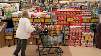 Una persona, vista de espaldas, empuja un carrito de compras lleno de comestibles en un pasillo de supermercado con estantes repletos de productos envasados