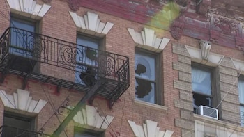 Primer plano de la fachada de un edificio de ladrillos con ventanas, una de ellas con el cristal roto, una escalera de incendios y un aire acondicionado