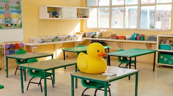 Aula con paredes amarillas, ventanas, varios pupitres y sillas verdes. Un pato de goma amarillo grande, un cuaderno y un lápiz se ven en un pupitre.