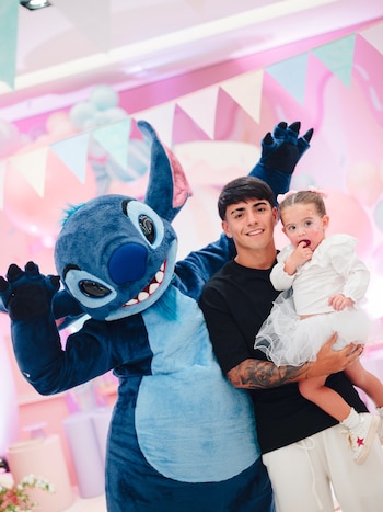 Un joven sostiene a una niña en brazos junto a un disfraz azul en un espacio decorado con guirnaldas de colores pastel