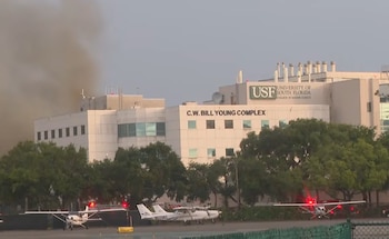 Edificio del complejo C.W. Bill Young de la USF con humo oscuro de la azotea. Varias avionetas y vehículos de emergencia con luces rojas en primer plano