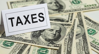 Una tarjeta blanca con la palabra "TAXES" en negro, apoyada sobre un conjunto desordenado de billetes de cien dólares americanos.