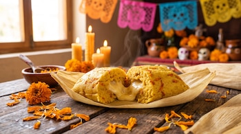 Un tamal abierto con queso derretido en una hoja de maíz sobre una mesa de madera con flores de cempasúchil, velas y papel picado al fondo.